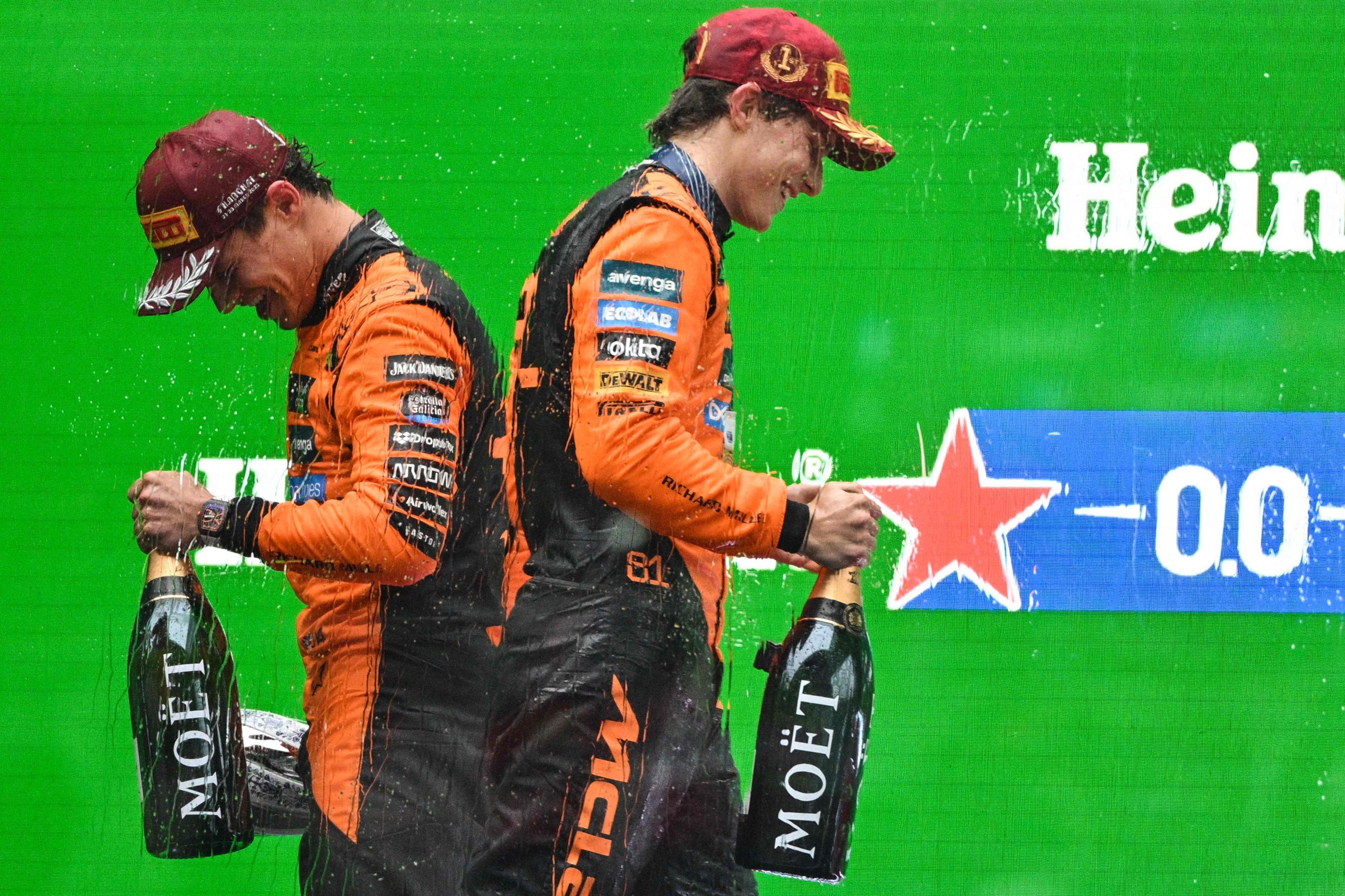 Two Formula 1 Mclaren drivers in orange and black uniforms celebrating with champagne bottles on a green background with Heineken branding.